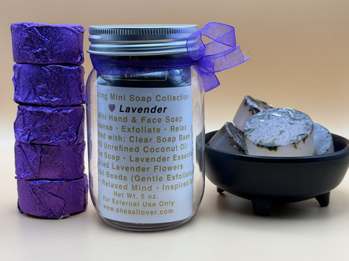 A lavender soap collection set featuring a jar of lavender hand and face soap with a blue lid and purple ribbon, a stack of three purple wrapped soap bars, and a small black bowl containing exfoliating lavender beads, all on a beige background.
